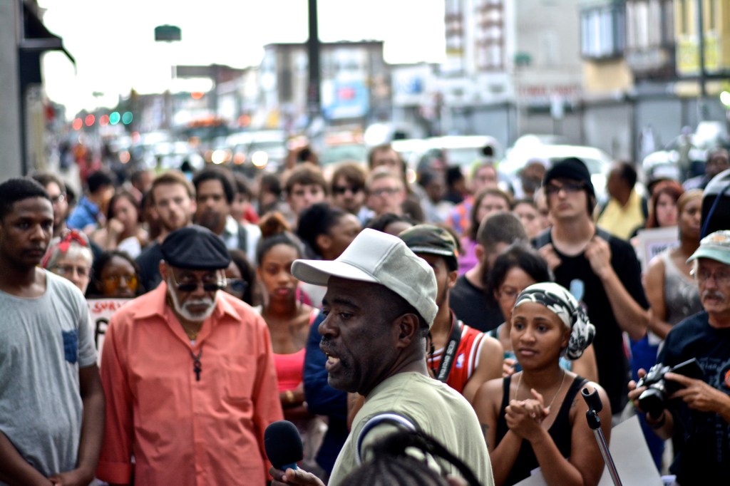 Crowd watches and listens to man as he tells his story of police brutality. 